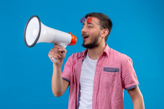 Young handsome traveler guy speaking to megaphone with happy face standing over blue background