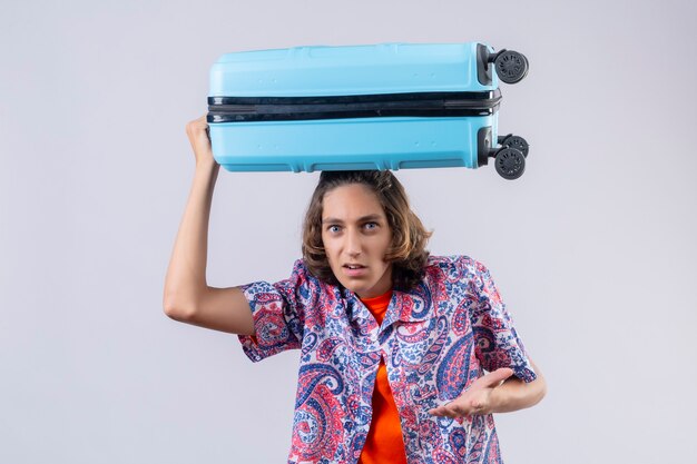 Free photo young handsome traveler guy standing with suitcase on head looking scared and disappointed over white background