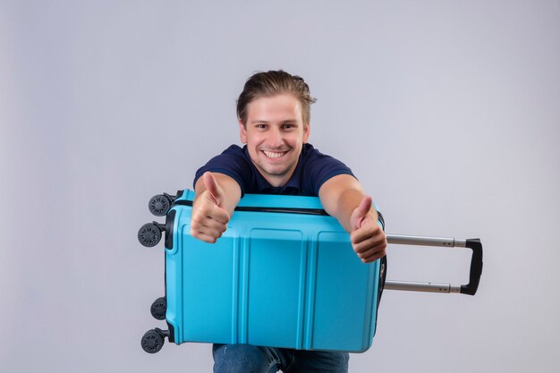 Young handsome traveler guy standing with suitcase looking at camera with happy face smiling cheerfully showing thumbs up over white background