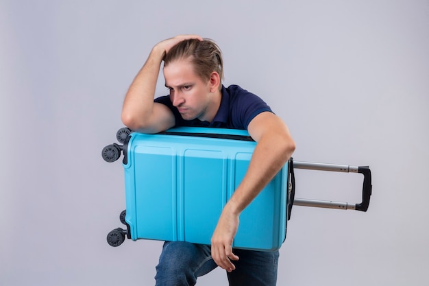 Ragazzo giovane viaggiatore bello in piedi con la valigia stressante tenendo le mani sulla testa stanco e frustrato su sfondo bianco