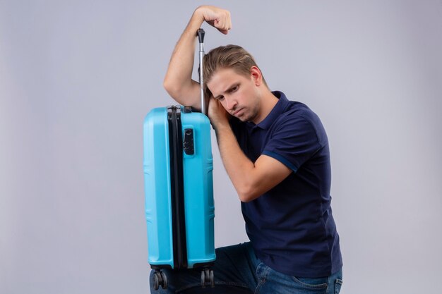 Young handsome traveler guy standing with suitcase with sad expression on face over white background