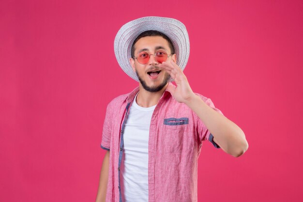 Young handsome traveler guy in summer hat wearing sunglasses calling someone with hand near mouth standing over pink background