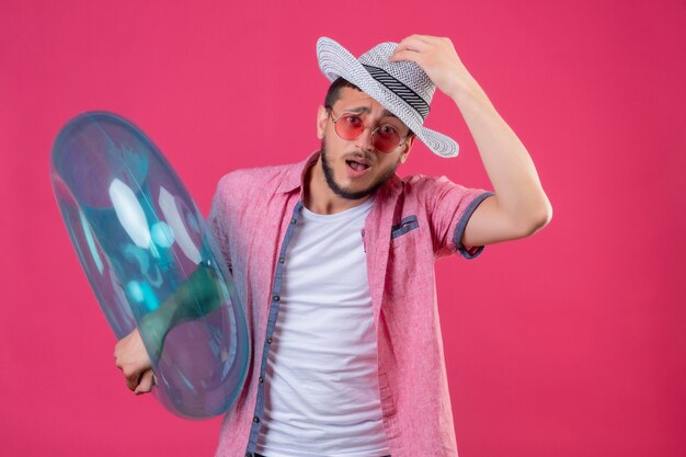 Young handsome traveler guy in summer hat wearing sunglasses holding inflatable ring clueless and confused touching his hat having no answer standing over pink background