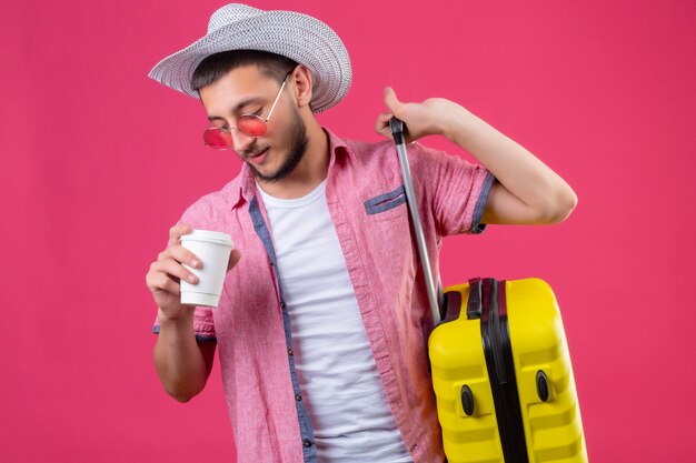 Young handsome traveler guy in summer hat wearing sunglasses holding suitcase and cup of coffee looking confident standing over pink background