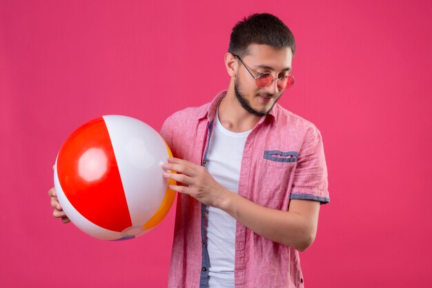 Молодой красивый путешественник в солнцезащитных очках держит надувной мяч, глядя в сторону с уверенной улыбкой на лице, стоящем на розовом фоне