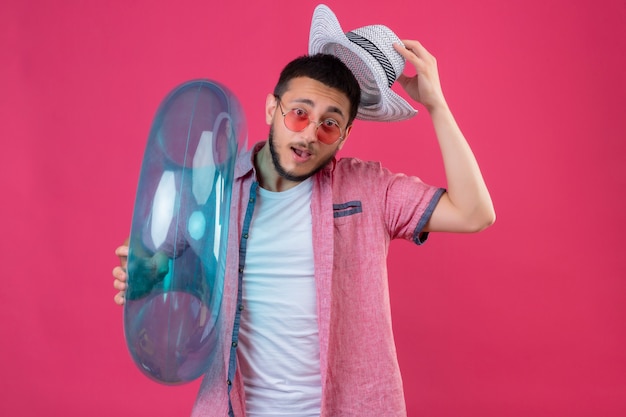 Young handsome traveler guy wearing sunglasses holding inflatable ring looking at camera amazed and surprised putting off his summer hat standing over pink background