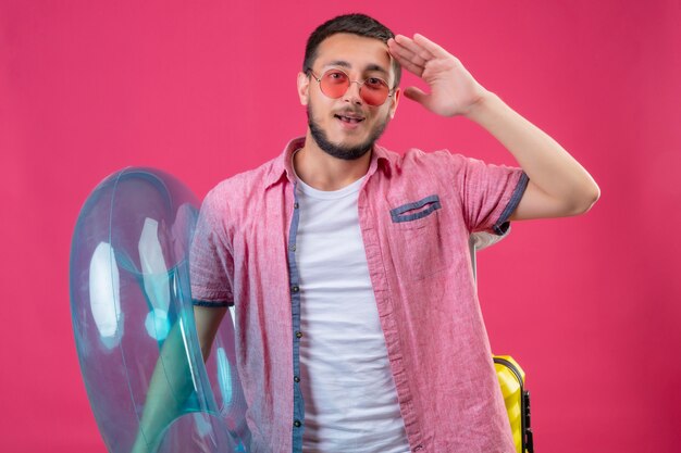 Young handsome traveler guy wearing sunglasses holding inflatable ring looking at camera positive and happy saluting standing over pink background