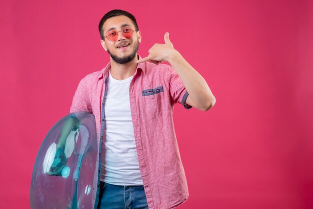 Young handsome traveler guy wearing sunglasses holding inflatable ring looking at camera smiling cheerfully making call me gesture standing over pink background