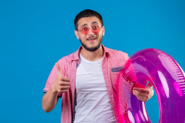 Young handsome traveler guy wearing sunglasses holding inflatable ring looking joyful and happy smiling cheerfully showing thumbs up standing over blue backgound