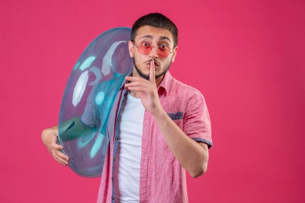 Young handsome traveler guy wearing sunglasses holding inflatable ring looking surprised making silence gesture with finger on lips standing over pink background