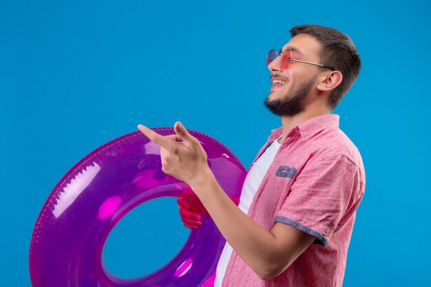 Young handsome traveler guy wearing sunglasses holding inflatable ring standing sideways smiling cheerfully pointing with finger to something over blue background