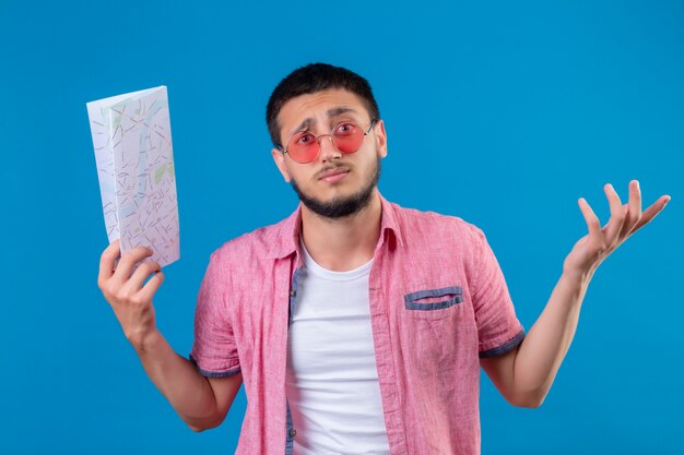 Young handsome traveler guy wearing sunglasses holding map clueless and confused with arms raised having no answer standing over blue background