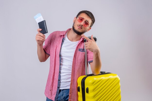 Young handsome traveler guy wearing sunglasses holding suitcase and air tickets looking at camera with sad expression on face standing over white background