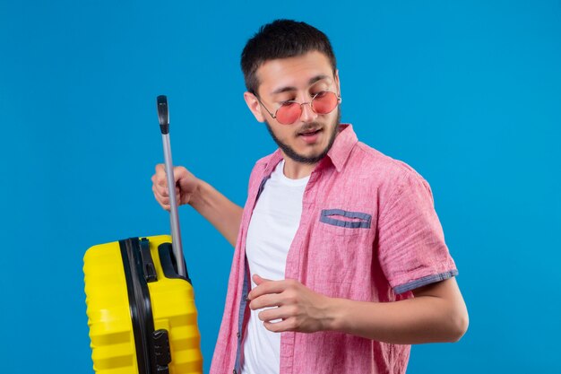 Young handsome traveler guy wearing sunglasses holding suitcase looking aside with confident expression standing over blue background