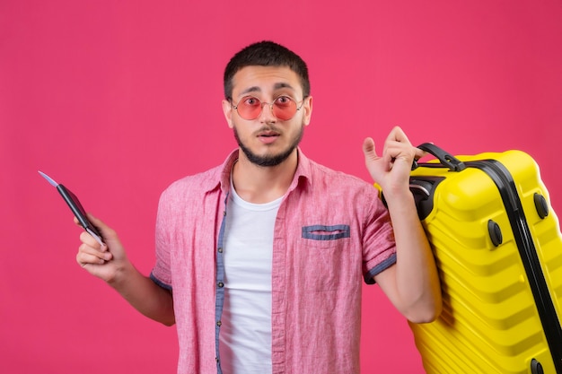 Young handsome traveler guy wearing sunglasses holding suitcase and tickets looking at camera with confuse expression standing over pink background