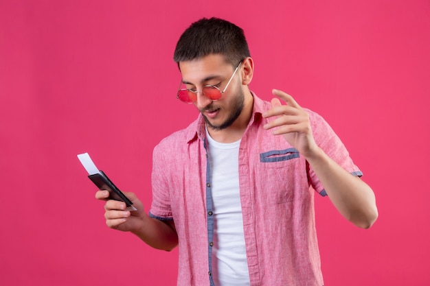 Young handsome traveler guy wearing sunglasses holding tickets looking aside with confident expression standing over pink background