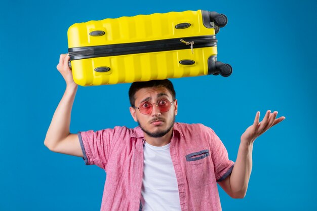 Young handsome traveler guy wearing sunglasses standing with suitcase on head clueless and confused standing with arms raised over blue background