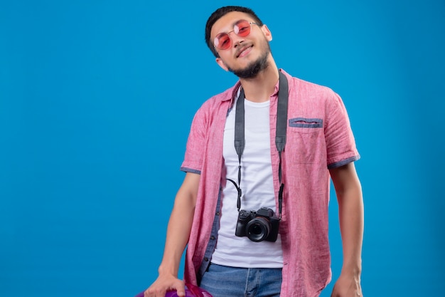 Young handsome traveler guy with camera wearing sunglasses looking at camera with confident smile standing over blue background