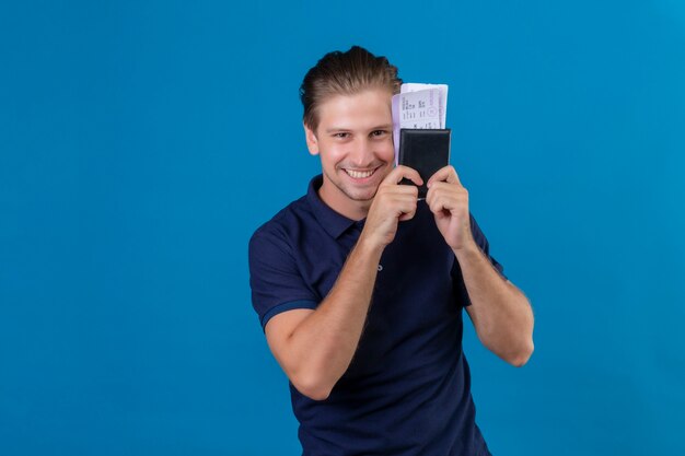 Young handsome traveler man holding air tickets happy and positive looking at camera with big smile on face standing over blue background