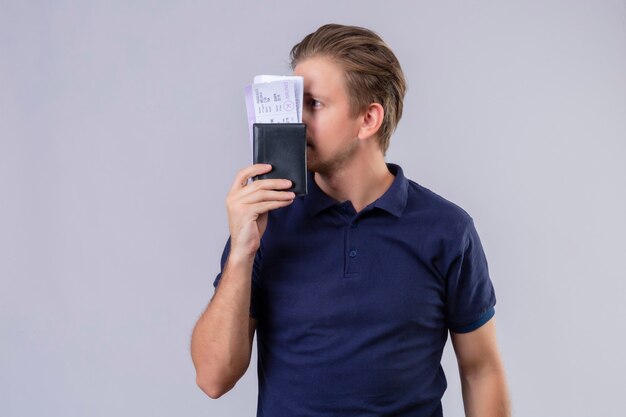 Young handsome traveler man holding air tickets hiding behind tickets standing over white background