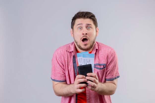 Young handsome traveler man holding airplane tickets surprised and amazed standing over white wall