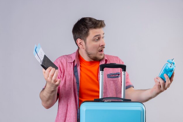 Молодой красивый путешественник мужчина держит синий чемодан и билеты на самолет, глядя на будильник, смущенный и разочарованный, стоящий над белой стеной