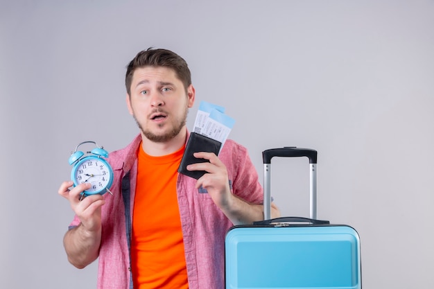 Young handsome traveler man holding blue suitcase and airplane tickets with alarm clock confused and disappointed standing over white wall