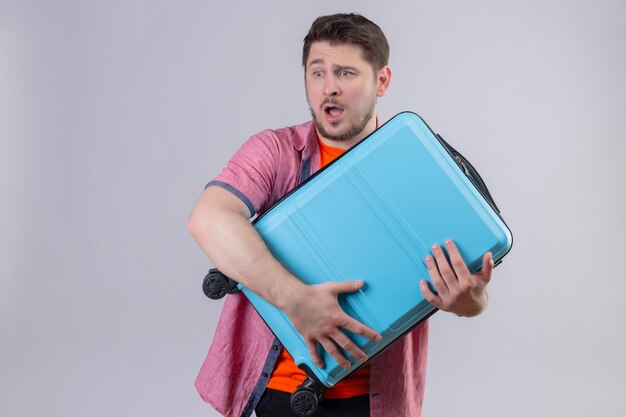 Young handsome traveler man holding blue suitcase looking aside surprised and amazed standing over white wall