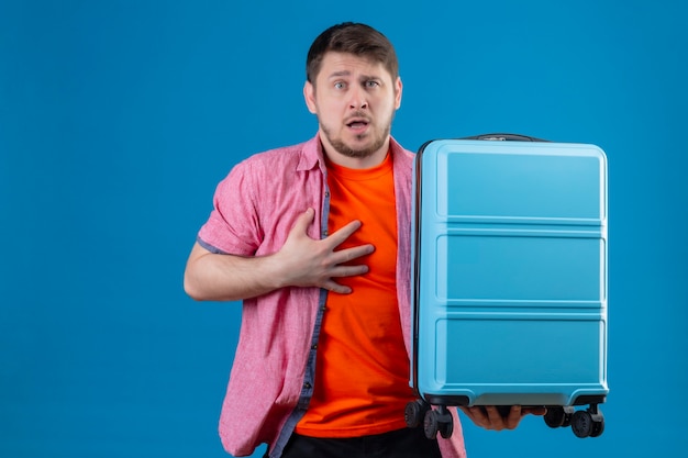 Молодой красивый путешественник мужчина держит чемодан эмоциональный и обеспокоенный с выражением страха на лице, стоящем над синей стеной