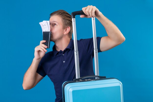 Young handsome traveler man holding suitcase and kissing tickets in his hand looking happy standing over blue background