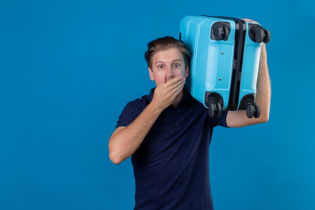 Молодой красивый путешественник мужчина держит чемодан, выглядящий изумленным и удивленным, прикрывая рот рукой, стоящей на синем фоне