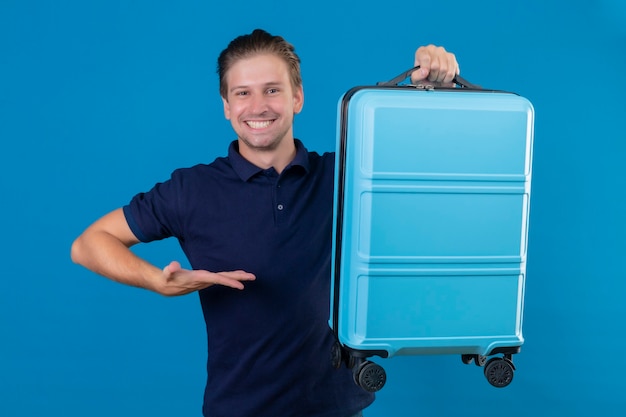 Uomo giovane viaggiatore bello che tiene la valigia guardando fiducioso presentando con il braccio della mano la sua valigia sorridendo allegramente in piedi su sfondo blu