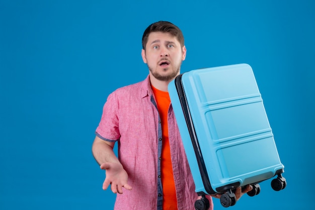 Free photo young handsome traveler man holding suitcase looking confused and very anxious standing over blue wall