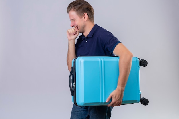 Young handsome traveler man holding suitcase smiling cheerfully with happy face standing sideways over white background