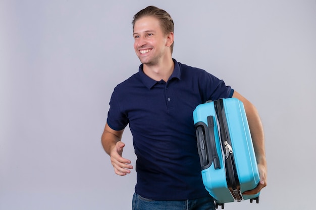 Free photo young handsome traveler man holding suitcase smiling cheerfully with happy face standing over white background