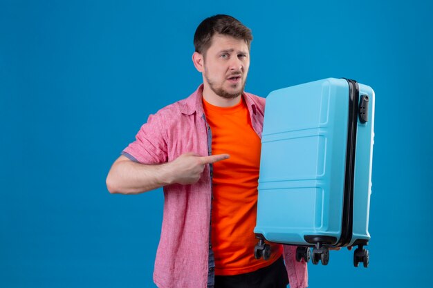 Young handsome traveler man holding suitcase with suspicious expression