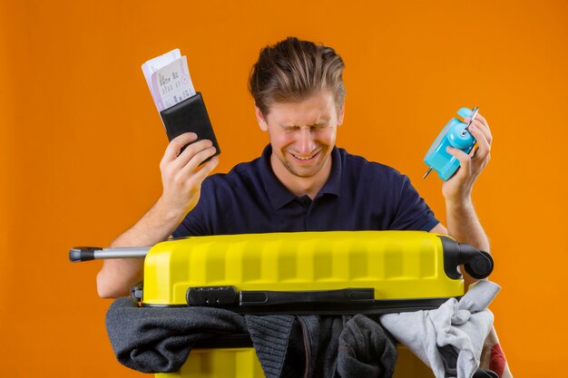 Young handsome traveler man standing with suitcase full of clothes holding alarm clock and air tickets with closed eyes unhappy crying over orange background