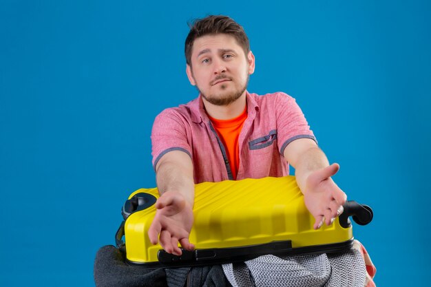 Uomo giovane viaggiatore bello in piedi con la valigia piena di vestiti, guardando confuso