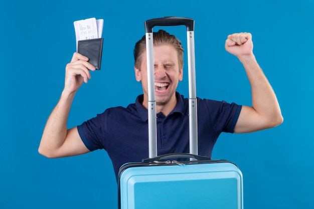 항공 티켓을 들고 가방으로 서 젊은 잘 생긴 여행자 남자는 파란색 배경 위에 유쾌하게 웃고 행복 한 얼굴로 종료
