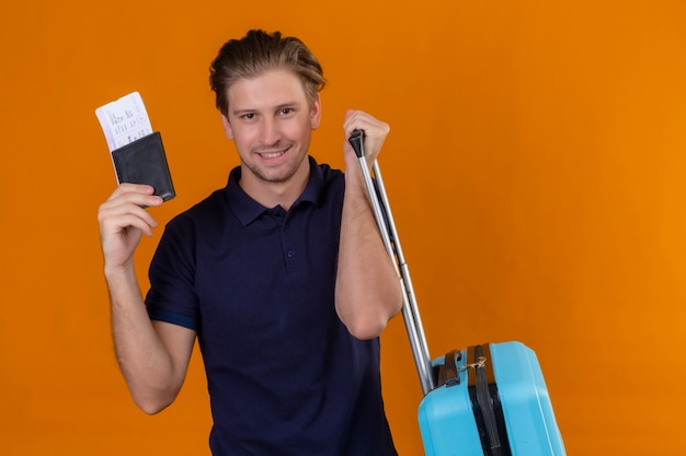 Young handsome traveler man standing with suitcase holding air tickets looking at camera with confident smile standing over orange background