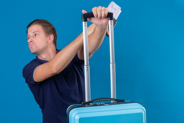 Young handsome traveler man standing with suitcase holding air tickets looking to the side displeased with sad expression on face over blue background