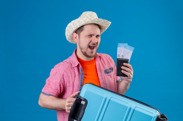 Young handsome traveler man in summer hat holding airplane tickets and suitcase very exited and happy standing over blue wall