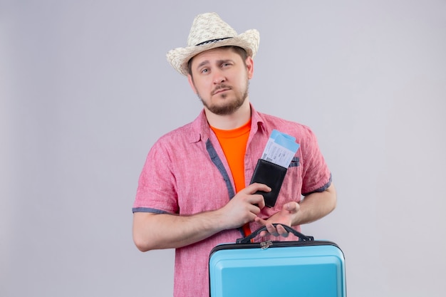 Young handsome traveler man in summer hat holding blue suitcase and airplane tickets with sad expression on face standing over white wall