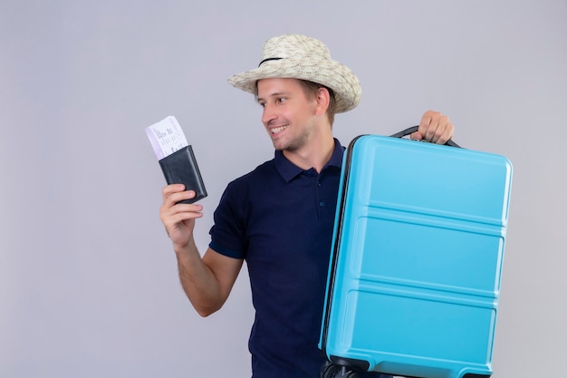 Young handsome traveler man in summer hat standing with suitcase holding air tickets looking aside with happy face smiling cheerfully over white background