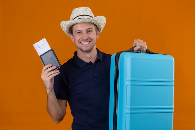 Foto gratuita uomo giovane viaggiatore bello in cappello estivo in piedi con la valigia in possesso di biglietti aerei che guarda l'obbiettivo con la faccia felice sorridente allegramente su sfondo arancione