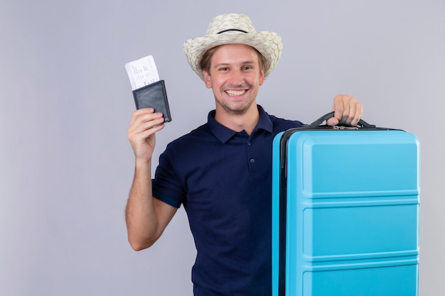 白い背景の上に元気に笑って幸せそうな顔でカメラを見て航空券を保持しているスーツケースを持って夏帽子立っている若いハンサムな旅行者男