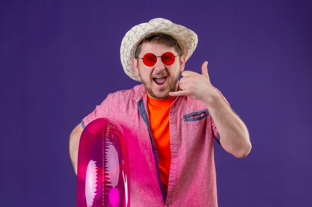 Young handsome traveler man in summer hat with inflatable ring making call me gesture