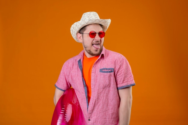 Free photo young handsome traveler man in summer hat with inflatable ring, sticking tongue out with disgusted expression