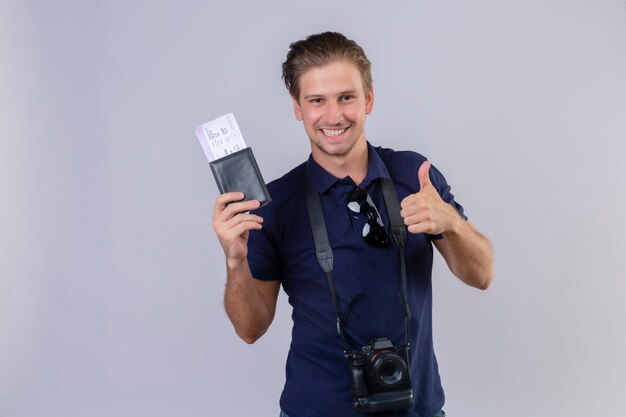 Молодой красивый путешественник с камерой, держащей авиабилеты, смотрит в камеру с улыбкой на лице, счастливым и позитивным, показывает палец вверх, стоя на белом фоне