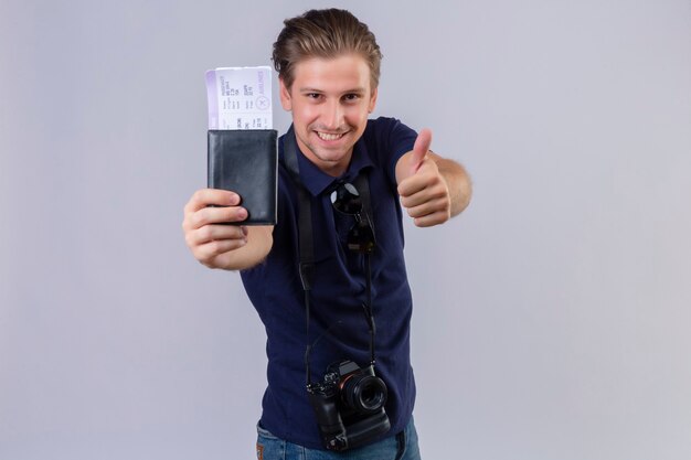 Free photo young handsome traveler man with camera holding air tickets looking at camera with smile on face happy and positive showing thumbs up standing over white background